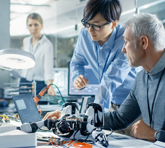 Two Engineers Talking while Working with Robot, Manipulating its Responses and Checking Data with Laptop Computer  In the Background Robotics Research Center Laboratory with Specialists Working 